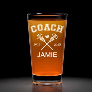 May include: A clear pint glass filled with an orange liquid. The glass has the word "COACH" in a white, arched font, with crossed lacrosse sticks and a ball. Below, it reads "2024 2025" and "JAMIE" in white.