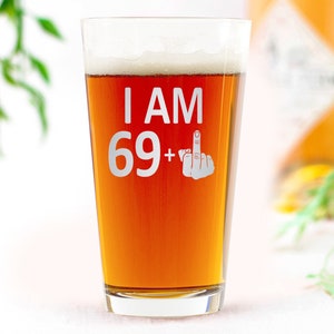 May include: A clear glass pint glass with a brown liquid inside. The glass has the text "I AM 69+" with a middle finger emoji etched on the side.