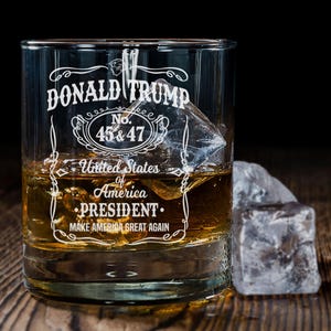 Peut inclure: Un verre &agrave; whisky en verre transparent avec un motif blanc qui dit "DONALD TRUMP No. 45 & 47 United States of America PRESIDENT MAKE AMERICA GREAT AGAIN". Le verre est rempli d'un liquide brun et de gla&ccedil;ons.
