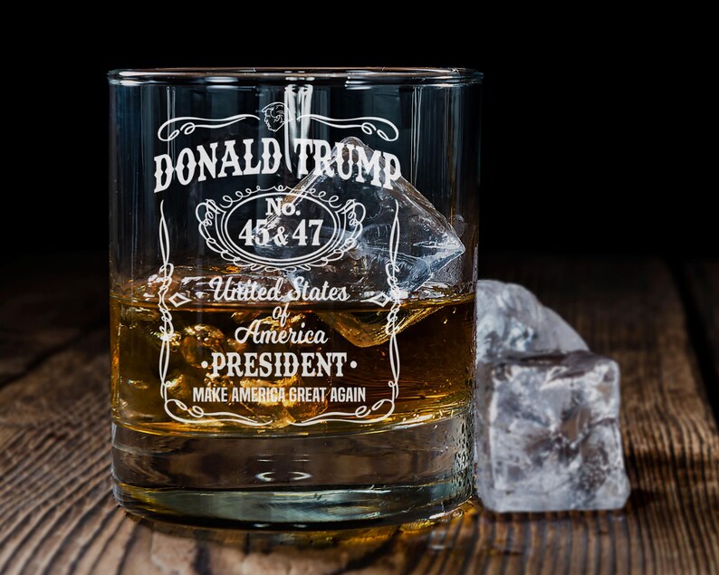 Peut inclure: Un verre &agrave; whisky en verre transparent avec un motif blanc qui dit "DONALD TRUMP No. 45 & 47 United States of America PRESIDENT MAKE AMERICA GREAT AGAIN". Le verre est rempli d'un liquide brun et de gla&ccedil;ons.