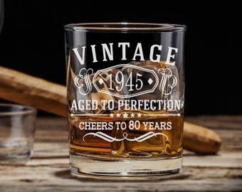 May include: A clear glass tumbler with the text "Vintage 1945 Aged to Perfection Cheers to 80 Years" etched on the side. The glass is filled with amber liquid and ice cubes.