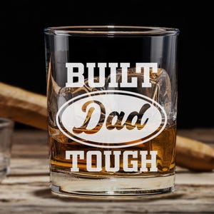 May include: A clear glass rocks glass with the text "BUILT TOUGH" and "Dad" in a white oval in the center. The glass is filled with amber liquid.