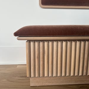 May include: A wooden bench with a brown velvet cushion and a fluted wood base. The bench has a rectangular backrest with a matching velvet insert. The wood has a natural finish, and the bench sits on a wooden floor.