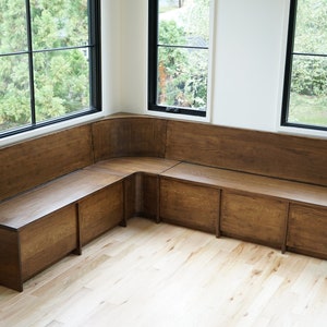 May include: A corner bench with a wooden seat and back, built-in storage compartments, and a curved design. The bench is positioned in front of a window with a view of trees.