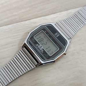 May include: A silver digital wristwatch with a black face and a silver metal strap. The watch face displays the time as 10:57:33. The watch also has a date display and a stopwatch function.