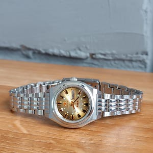 Può includere: Un orologio Orient argentato con quadrante dorato e cinturino argentato. Il quadrante mostra il giorno della settimana e il logo Orient. L'orologio è appoggiato su una superficie di legno.