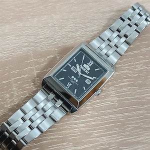 Puede incluir: Un reloj Orient plateado con una esfera negra y una correa de metal plateado. La esfera del reloj tiene agujas y marcadores plateados, y la palabra "Orient" está impresa en blanco en la esfera.