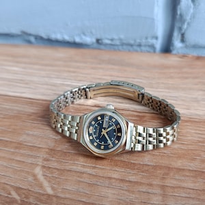 Può includere: Un orologio da polso vintage color oro con quadrante nero e lancette dorate. L'orologio presenta un datario e un cinturino in metallo a maglie. L'orologio è appoggiato su una superficie di legno.