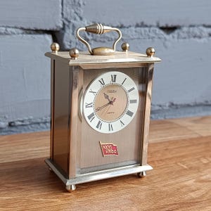 May include: A vintage brass-toned clock with a rectangular frame and a handle. The clock face features Roman numerals and a red emblem. The clock is set on a wooden surface.