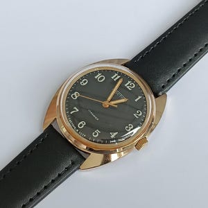 Op de afbeelding: Een gouden polshorloge met een zwarte wijzerplaat en een zwarte leren band. De wijzerplaat van het horloge heeft witte Arabische cijfers en een kleine secondewijzer. Het horloge is gelabeld met "СССР" en "17 jewels".