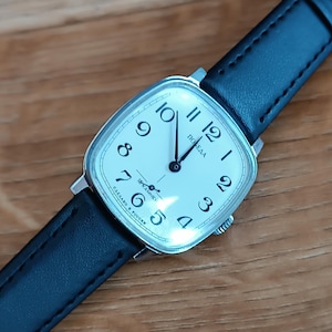 Puede incluir: Un reloj de pulsera vintage con una esfera cuadrada de color crema y números arábigos negros. El reloj tiene una caja plateada y una correa de cuero negro. La esfera del reloj tiene la palabra "ПОБЕДА" y otro texto. El reloj está sobre una superficie de madera.