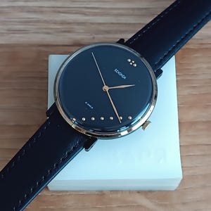 Puede incluir: Un reloj de pulsera Sekonda con esfera negra y manecillas y detalles dorados. El reloj tiene una correa de cuero negro y se muestra sobre un bloque rectangular blanco. La esfera del reloj tiene el texto "SEKONDA" y "21 JEWELS".