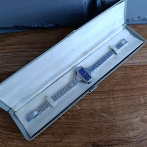 May include: A vintage silver-toned wristwatch with a rectangular blue face, presented in a light gray case with a white interior. The watch has a linked band and is secured with white paper. The case is open, revealing the watch's design.