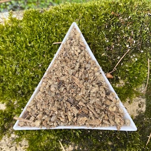 Puede incluir: Un plato blanco triangular lleno de una mezcla de hierbas marrones, secas y desmenuzadas. El plato está sentado sobre un lecho de musgo verde.