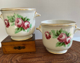 May include: Two white ceramic flower pots with gold trim and pink rose floral designs. The pots have handles on the sides. One pot is sitting on a wooden box.
