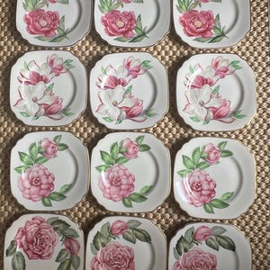 May include: A set of twelve square, white porcelain plates. Each plate features a different floral design in shades of pink and green, with gold trim. The floral patterns include peonies, magnolias, and roses.
