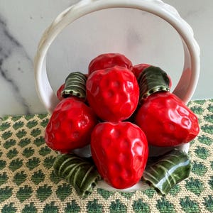 May include: A white ceramic basket filled with red and green ceramic strawberries. The basket has a white handle and is sitting on a green and white patterned surface.