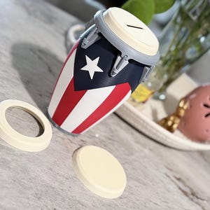 May include: A drum-shaped money box decorated with the Puerto Rican flag. The design includes a white star on a blue section, with red and white stripes. Two beige circular pieces are placed nearby. The bank is approximately 15 cm tall.