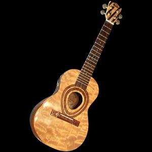 Puede incluir: Un ukelele marrón claro con un acabado brillante. El instrumento presenta una boca con círculos concéntricos y un clavijero a rayas. El cuerpo tiene un patrón de vetas de madera natural.