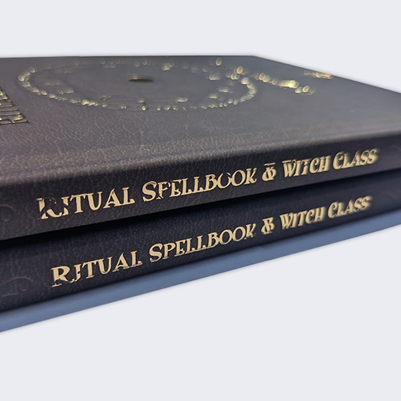 Op de afbeelding: Twee donkergrijze hardcover boeken met gouden letters op de rug. De tekst luidt "Ritual Spellbook of Witch Class".