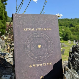 Op de afbeelding: Een donkerbruin, in leer gebonden boek met gouden letters waarop "Ritual Spellbook & Witch Class" staat. Het boek heeft een gouden cirkel met een pentagram en een gouden ster in het midden. Het boek ligt op een stenen muur met witte bloemen op de achtergrond.