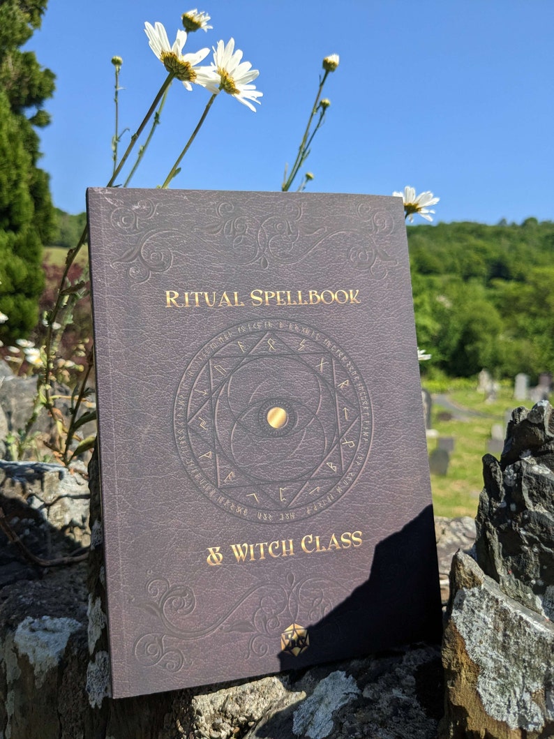 Op de afbeelding: Een donkerbruin, in leer gebonden boek met gouden letters waarop "Ritual Spellbook & Witch Class" staat. Het boek heeft een gouden cirkel met een pentagram en een gouden ster in het midden. Het boek ligt op een stenen muur met witte bloemen op de achtergrond.