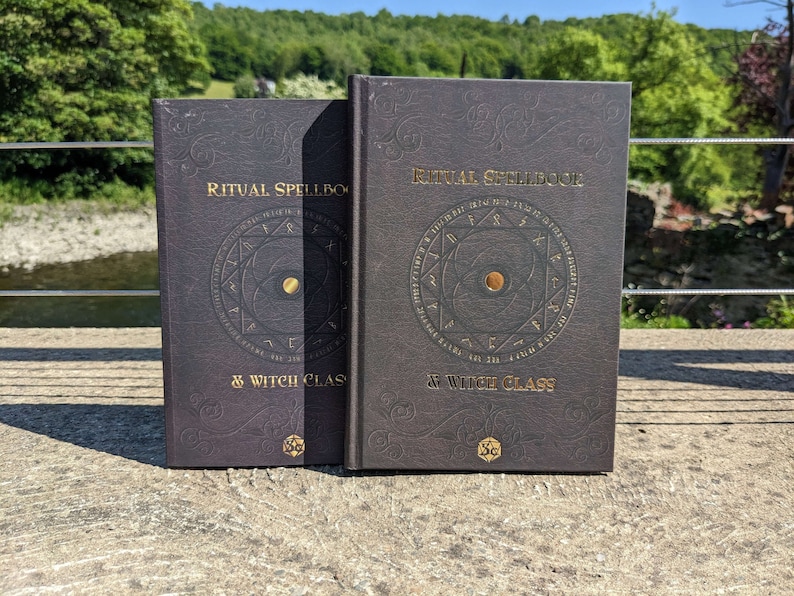 Op de afbeelding: Twee zwarte hardcover boeken met gouden letters en een gouden symbool op de voorkant. De boeken zijn getiteld "Ritual Spellbook & Witch Class".