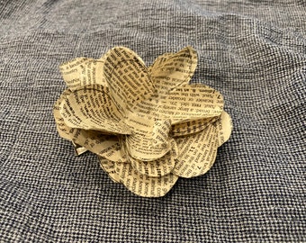 May include: A paper flower made from pages of a book. The flower is made of several layers of paper, each with text printed on it. The flower is a light brown color and has a slightly textured surface.