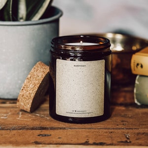 Puede incluir: Un tarro de vela de vidrio marrón con una tapa de corcho. La etiqueta en el tarro dice "HappySoy" y "Handgemacht" con un símbolo de corazón. El texto debajo dice "Duftkerze aus naturlichem Sojawachs".