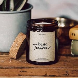 May include: A brown glass candle jar with a cork lid. The candle is labelled "HappySoy" and has the text "Beste Trainerin" on the front. The candle is surrounded by a wooden surface.