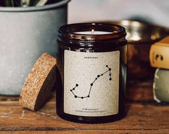 May include: A brown glass candle jar with a cork lid. The candle is labelled "HAPPYSOY" and features a black silhouette of the Sagittarius constellation. The text on the label reads "MIT ♥ HANDGEMACHT DUFTKERZE AUS NATÜRLICHEM SOJAWACHS".