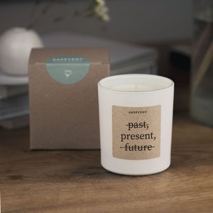 Könnte beinhalten: Eine weiße Kerze in einem Glas mit dem Text "past, present, future" auf einem braunen Etikett. Eine braune Schachtel mit einem grünen Etikett befindet sich hinter der Kerze. Die Kerze und die Schachtel stehen auf einer Holzoberfläche.