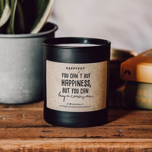 May include: Black soy candle with a brown paper label that reads "You can't buy happiness, but you can buy a caravan." The candle is on a wooden surface with a stack of books in the background.