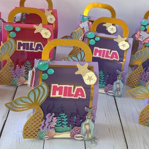May include: A collection of mermaid-themed gift bags in shades of pink and purple. Each bag features a gold handle, the name "MILA", a mermaid tail, and underwater-themed embellishments like seashells and starfish. The bags are decorated with gold accents.