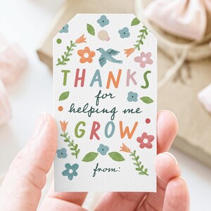 May include: A white gift tag with the words "THANKS for helping me GROW" in colorful letters. The tag is decorated with flowers, leaves, and a bird illustration. The tag also has the word "from:" at the bottom.