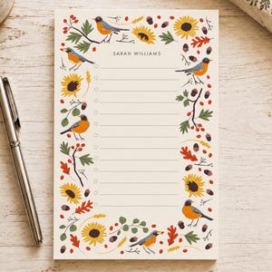 May include: A rectangular notepad with a cream background featuring a border of illustrated orange-breasted birds, yellow sunflowers, brown acorns, and red berries. The center contains horizontal lines with circular checkboxes and the name SARAH WILLIAMS.