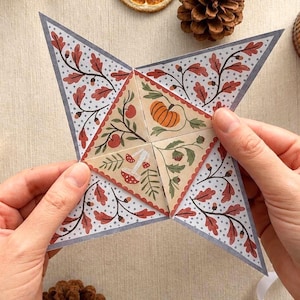 Pode incluir: Um objeto de papel em forma de estrela de quatro pontas com um quadrado central dividido em quatro triângulos. As pontas externas têm um fundo branco com folhas de carvalho vermelhas e bolotas. O centro mostra ilustrações de abóbora e cogumelos.