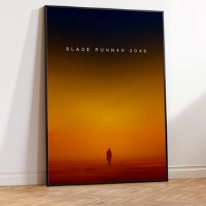 May include: A black framed poster with a minimalist design featuring a silhouette of a person walking towards a bright orange and yellow sunset. The text "BLADE RUNNER 2049" is printed in white at the top of the poster.