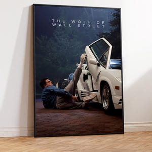May include: A framed poster for "The Wolf of Wall Street." The image depicts a white sports car with an open door and a person lying on the ground beside it. The poster is encased in a black frame.
