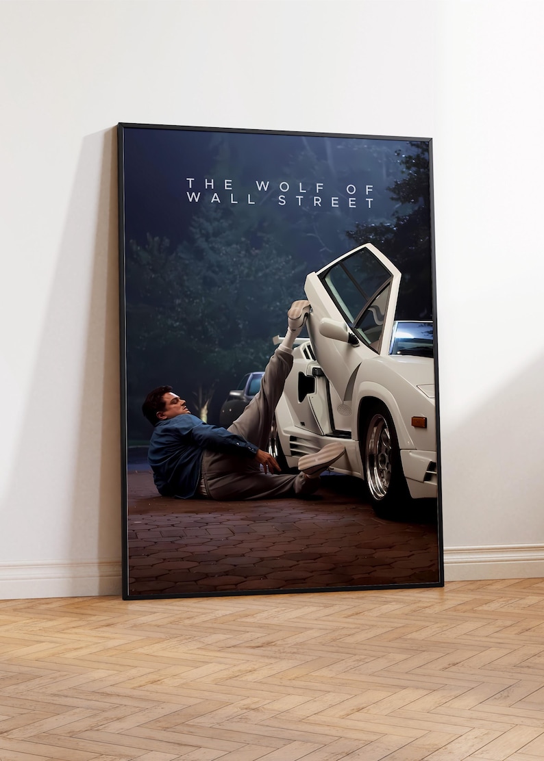 May include: A framed poster for "The Wolf of Wall Street." The image depicts a white sports car with an open door and a person lying on the ground beside it. The poster is encased in a black frame.