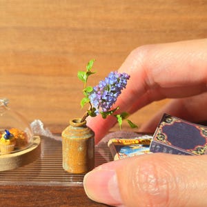 Peut inclure: Une scène miniature comprenant un petit vase en céramique marron contenant un brin de lilas violet, une cloche en verre transparent recouvrant trois minuscules cupcakes sur un socle en bois, et un petit livre rectangulaire à couverture bleu foncé.