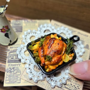 Op de afbeelding: Miniatuur gebraden kip in een zwarte vierkante pan met groenten en kruiden. Het gerecht staat op een wit kanten kleedje en krant. Een kleine witte kan met bloemen staat op de achtergrond.