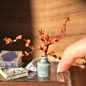 May include: Miniature scene featuring a small gray vase holding a branch with red flowers. Two tiny books with blue and beige covers are nearby. The scene is set on a glass surface with a wooden background.