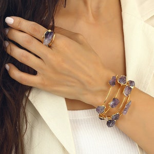 May include: A gold bangle bracelet with multiple bands adorned with purple amethyst stones. The bracelet is worn on a person's wrist.