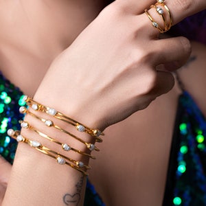 May include: A set of five gold bangles with white pearl accents. The bangles are stacked on a person's wrist.