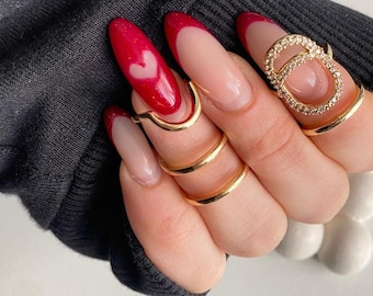 May include: Close-up of a hand with long, almond-shaped nails painted with a red and white ombre design. The nails feature a heart design on the ring finger. The hand is wearing several gold rings, including one with a large, circular design.
