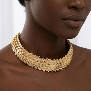 Könnte beinhalten: Eine goldfarbene Statement-Halskette mit einem strukturierten, geschichteten Design. Die Halskette wird um den Hals getragen und hat ein detailliertes, blattartiges Muster. Der Schmuck ist ein auffälliges Accessoire.