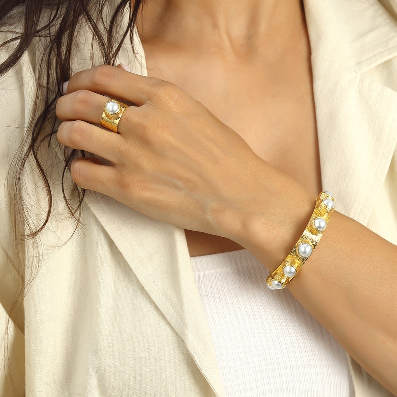May include: A gold-toned ring and bracelet set with faux pearls. The ring is wide and has a single pearl in the center. The bracelet is a bangle style with multiple pearls around the circumference.
