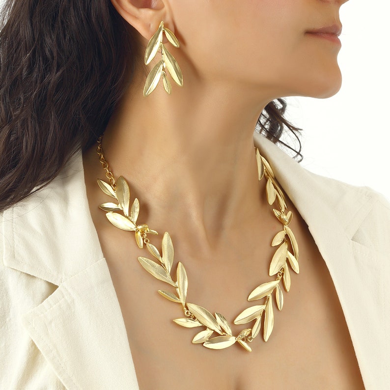 May include: A gold necklace and earrings set featuring a design of olive branches. The necklace is a statement piece with large, detailed leaves.