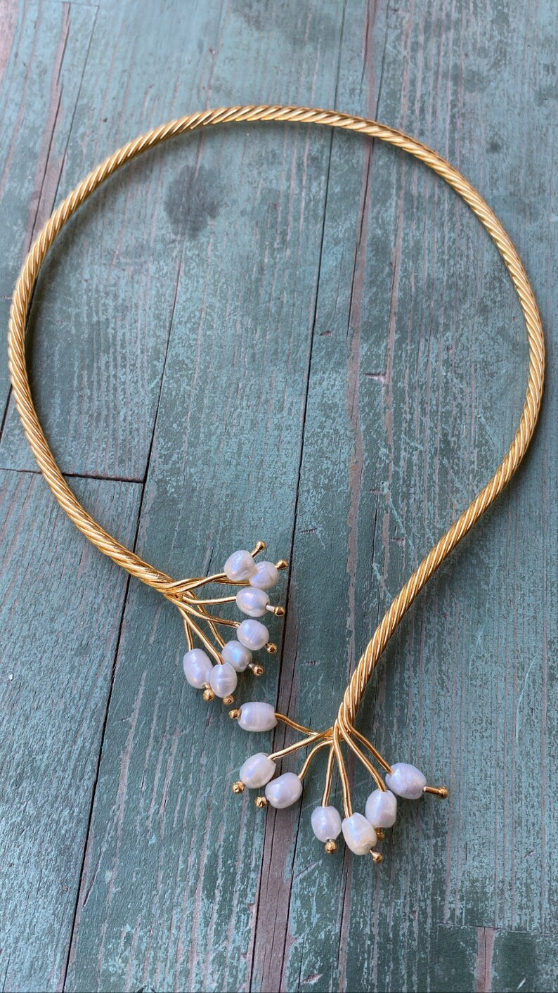 May include: A gold choker necklace with a twisted rope design and pearl accents. The pearls are clustered at the ends of the necklace, creating a delicate and feminine look.